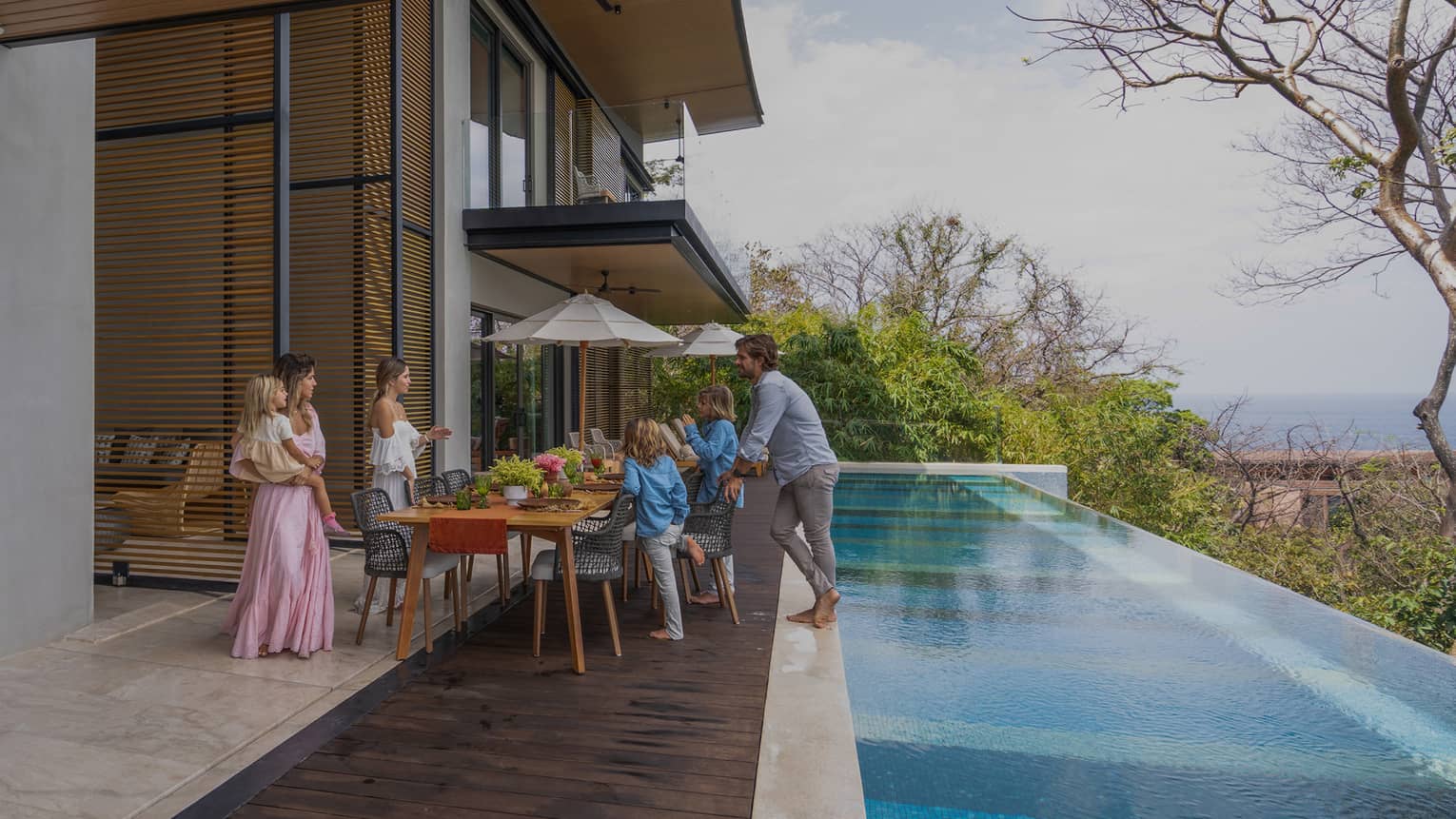 Family gathers around dining table set outside on a deck next to a private infinity-edge pool