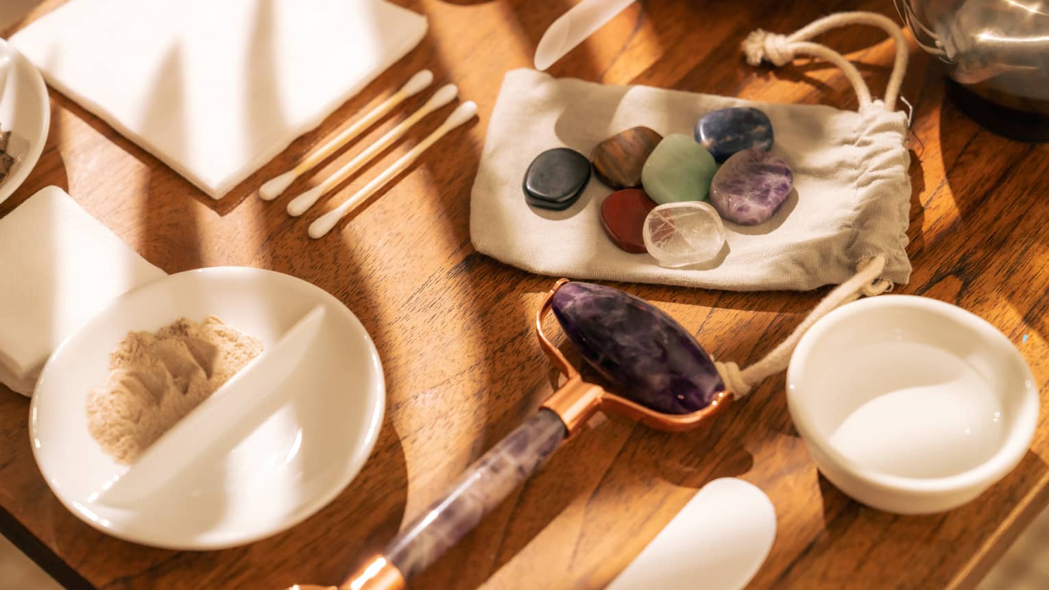 Face roller, gemstones, cotton swabs and a variety of small white bowls and other instruments sit on a sun-lit wooden table