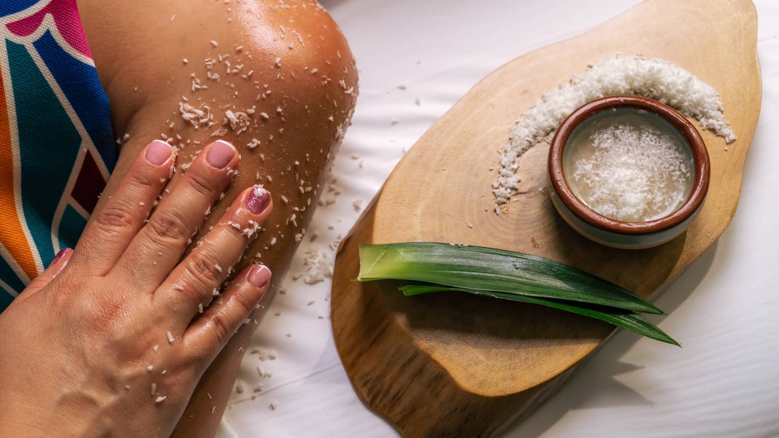 Small bowl of sugar scrub sits next to two green leaves on a wooden platter as a hand rubs the scrub on a person's shoulder