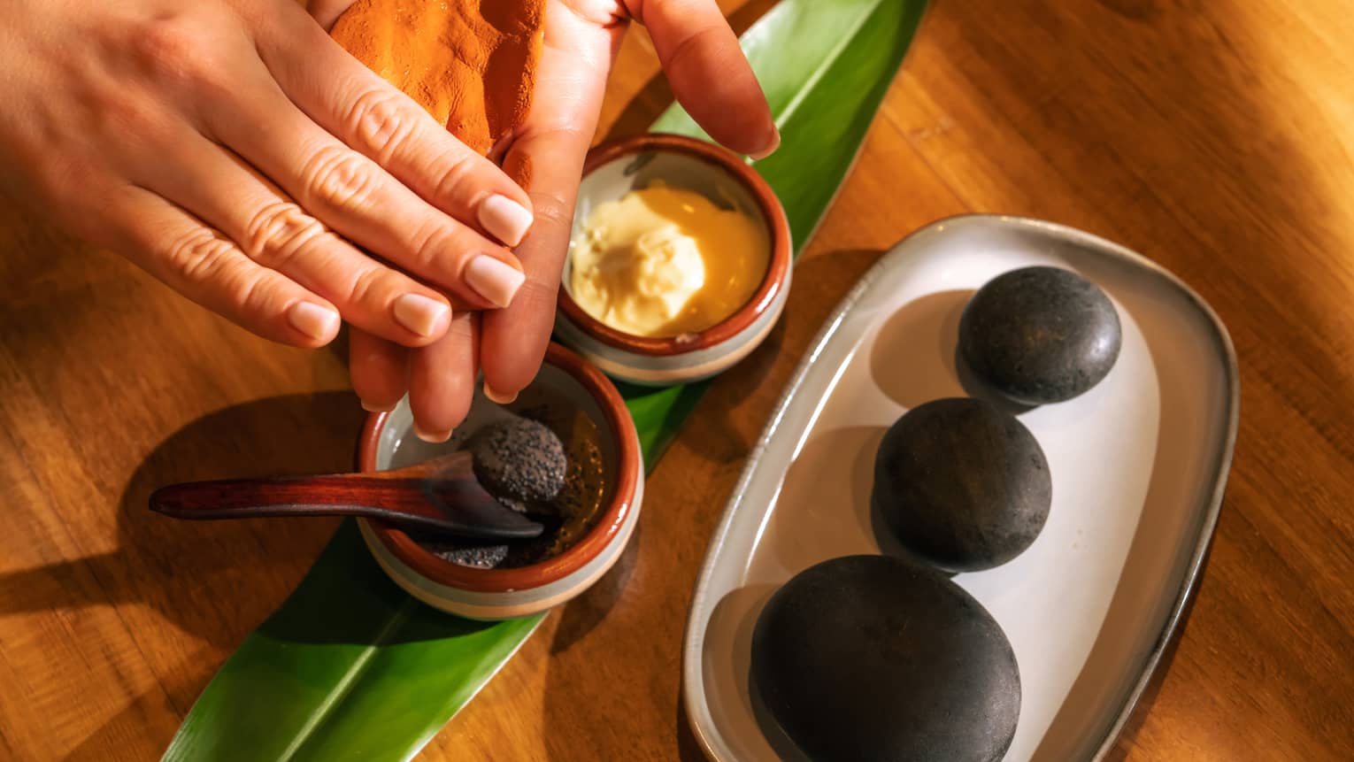 Pair of hands tests spa ingredients from two small stoneware bowls set on a fresh green leaf with three black stones placed on a dish nearby