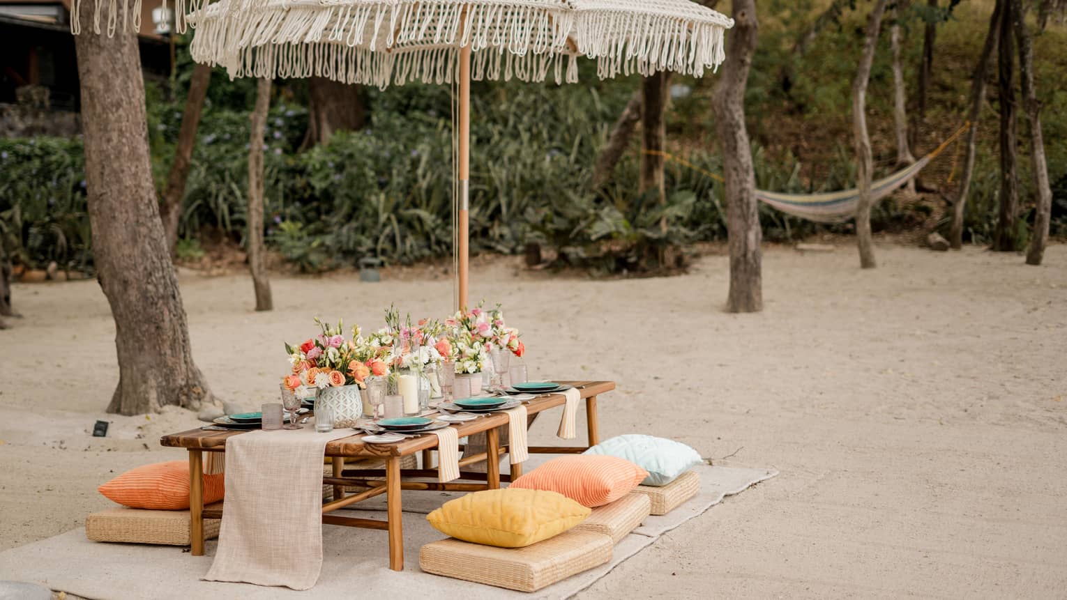 Wooden dining table set up with floral centrepieces and candles beneath an umbrella on a beach