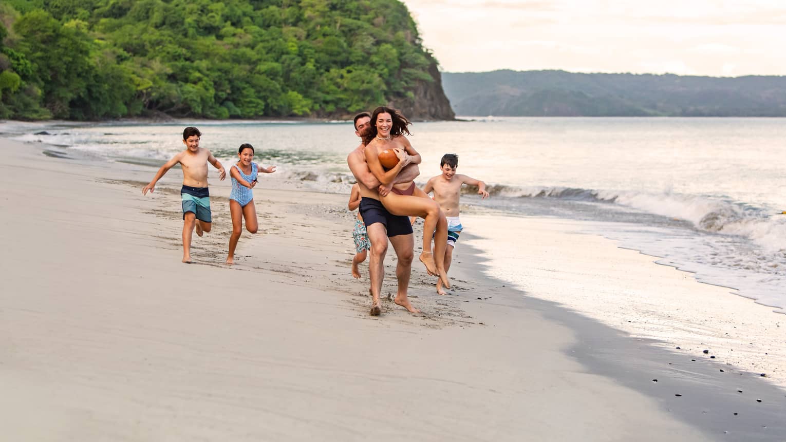 Family of six runs along the waterline on a sandy beach with a tree-covered outcropping in the background
