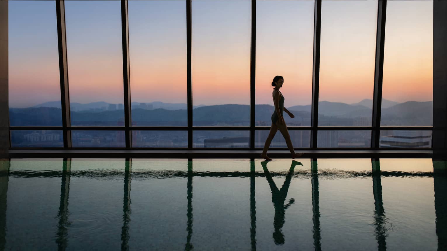 A guest in silhouette walks along the edge of a glassy indoor pool beside a wall of windows with mountain views at sunset.