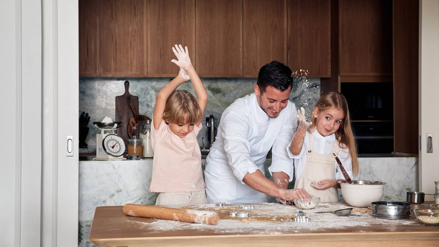 Chef wearing white chef's jacket helps to young children make pizza dough on a kitchen island