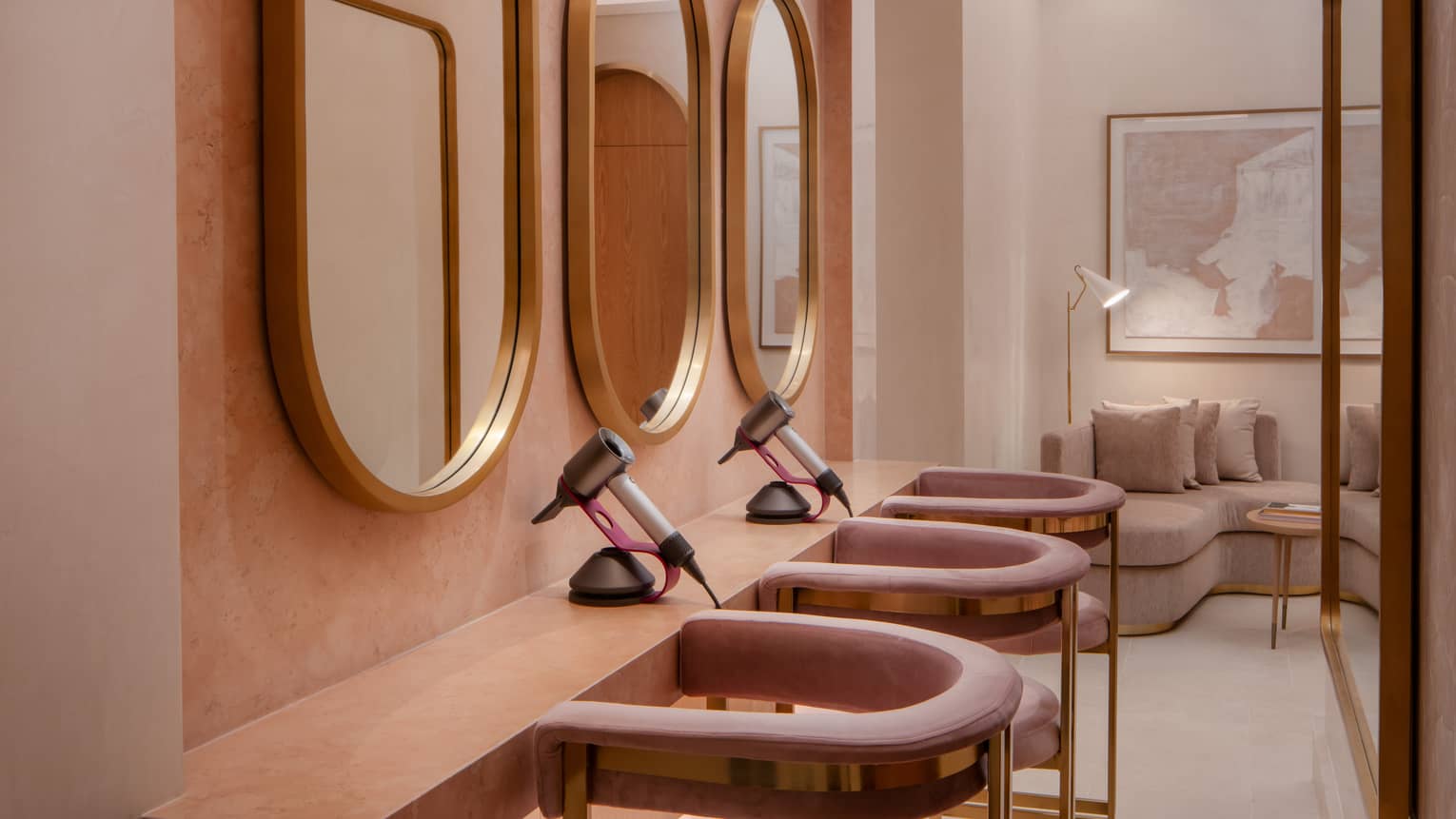 Dressing room with three gold oval mirrors on a wall in front of a small counter with two blow driers set on top and three pink curved-arm chairs pulled up to it