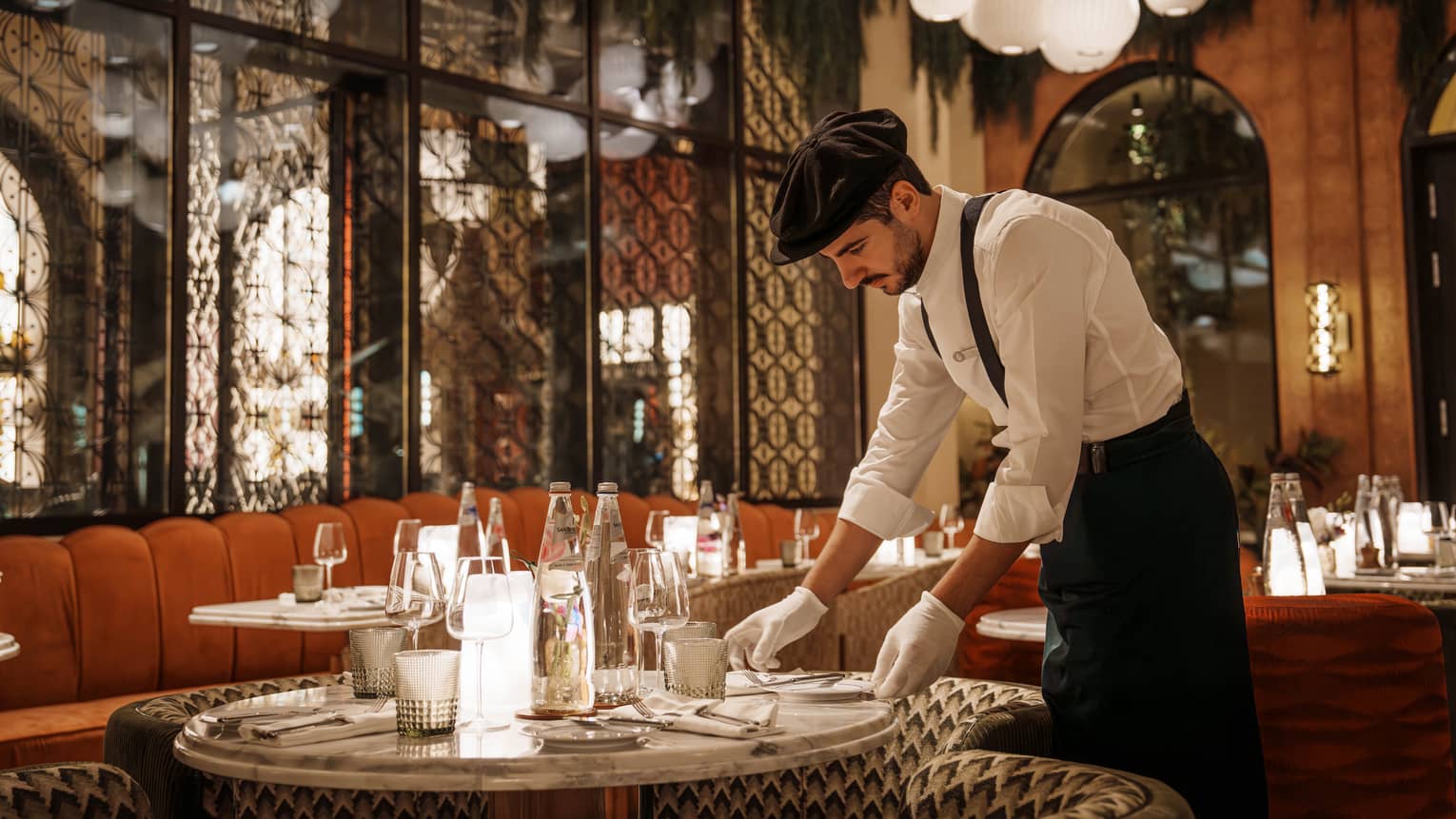 Waiter wearing white button-down shirt, black pants, black suspenders, black beret and white gloves arranges a plate on a table in an Art Deco?style restaurant
