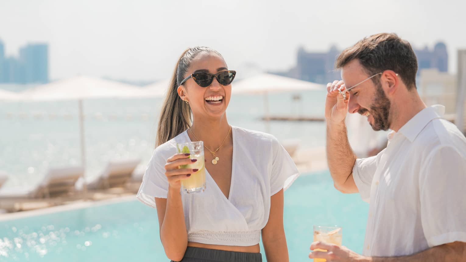 Two people wearing white shirts and sunglasses stand smiling and laughing next to a pool, each holding a drink in their hand