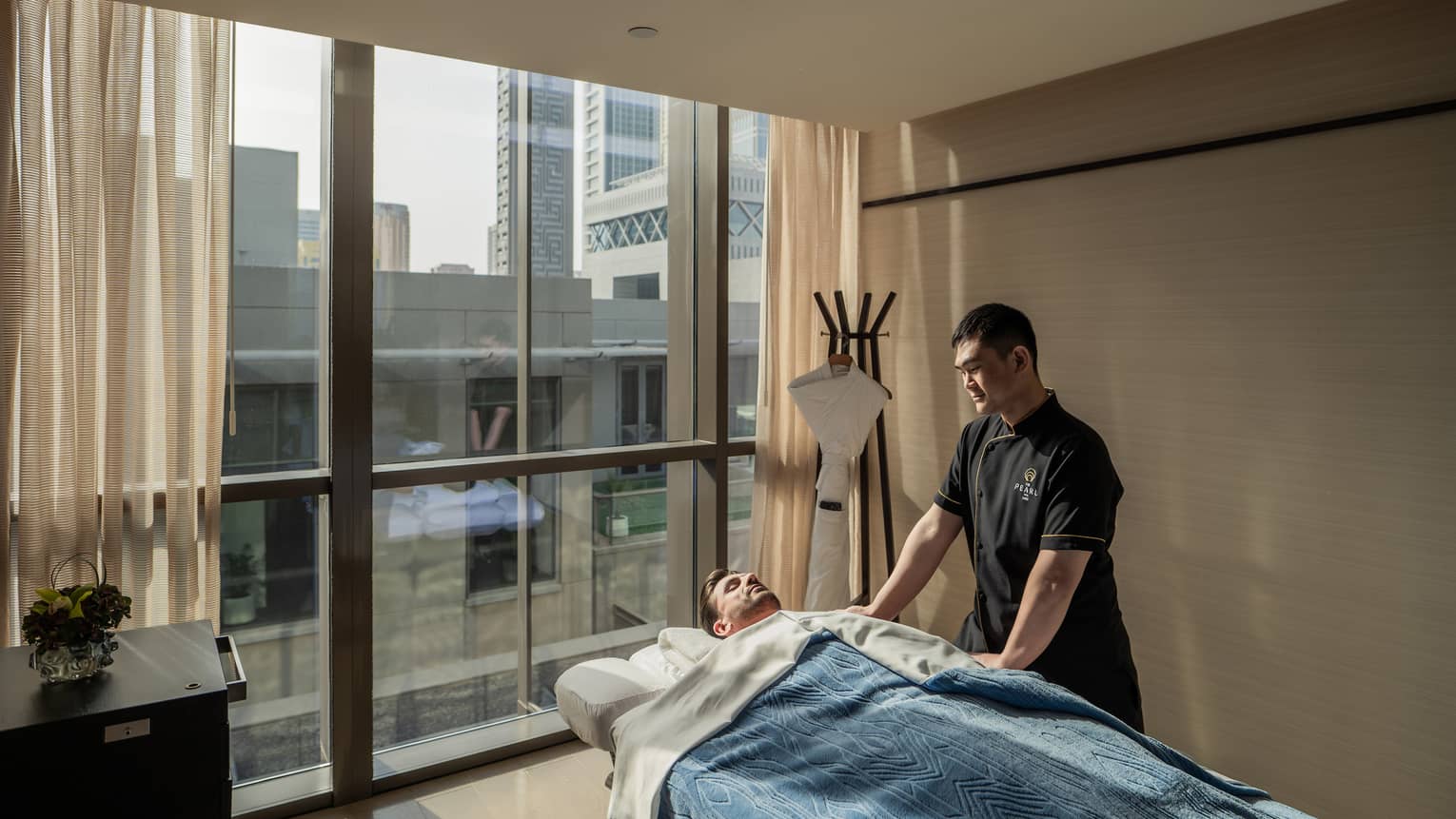 Male spa attendant stands next to man laying on spa treatment bed covered in a blue blanket