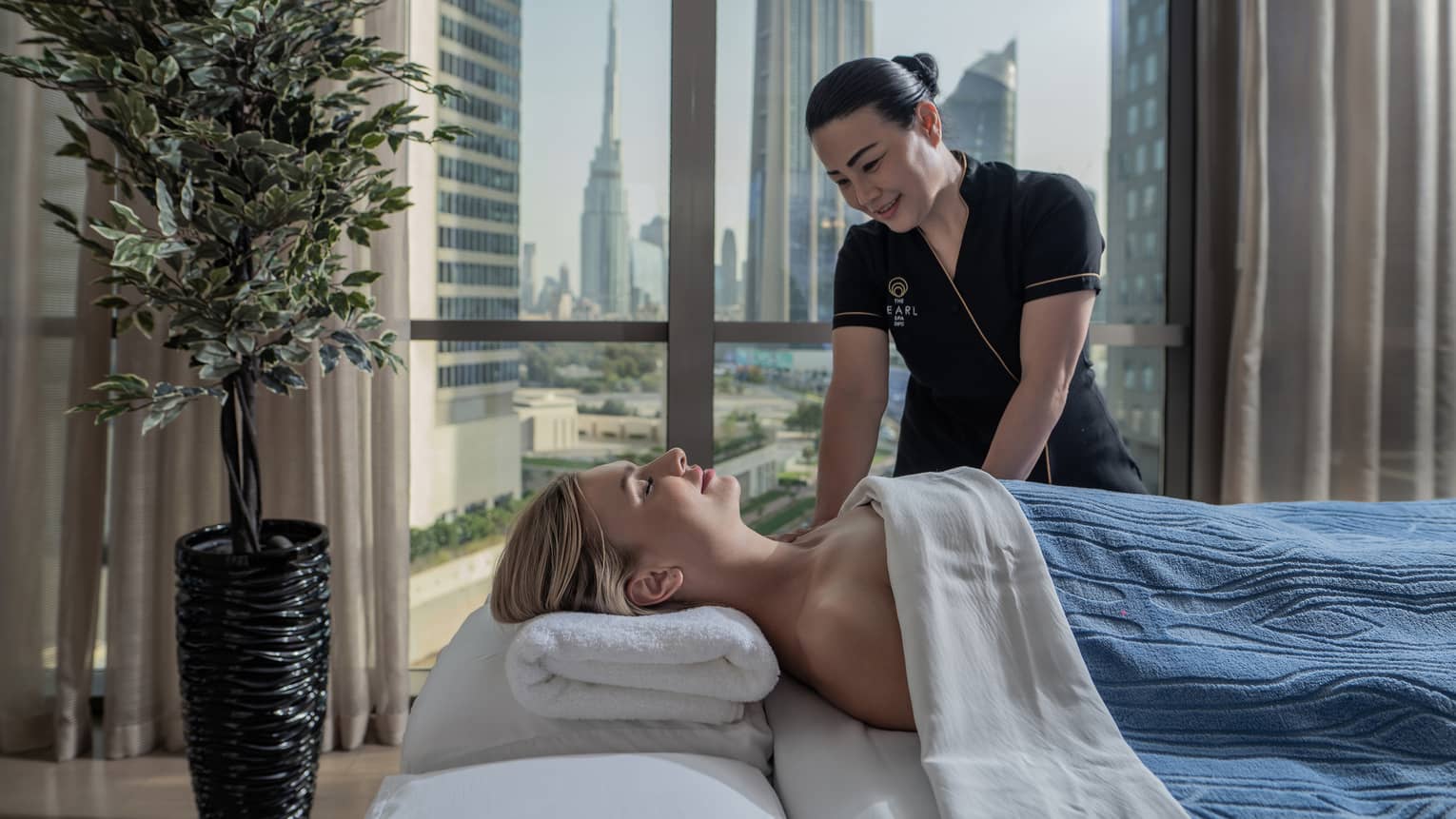 A guest enjoys a massage from a Hotel employee. Behind them, a window frames tall glass skyscrapers and clusters of trees.