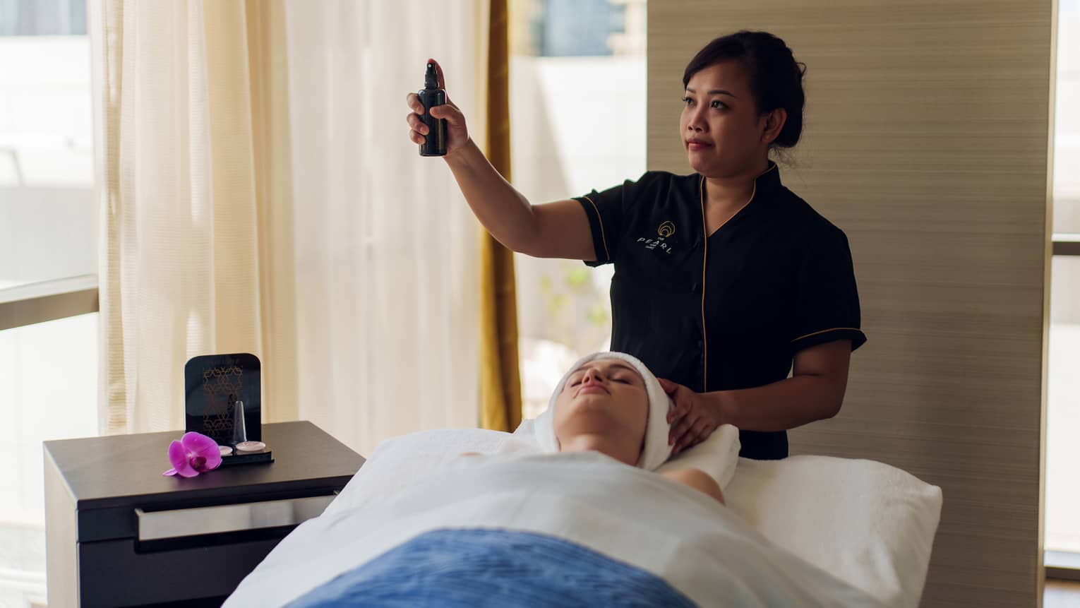 Spa therapist wearing black short-sleeve shirt stands behind person laying on a massage table covered in a white blanket