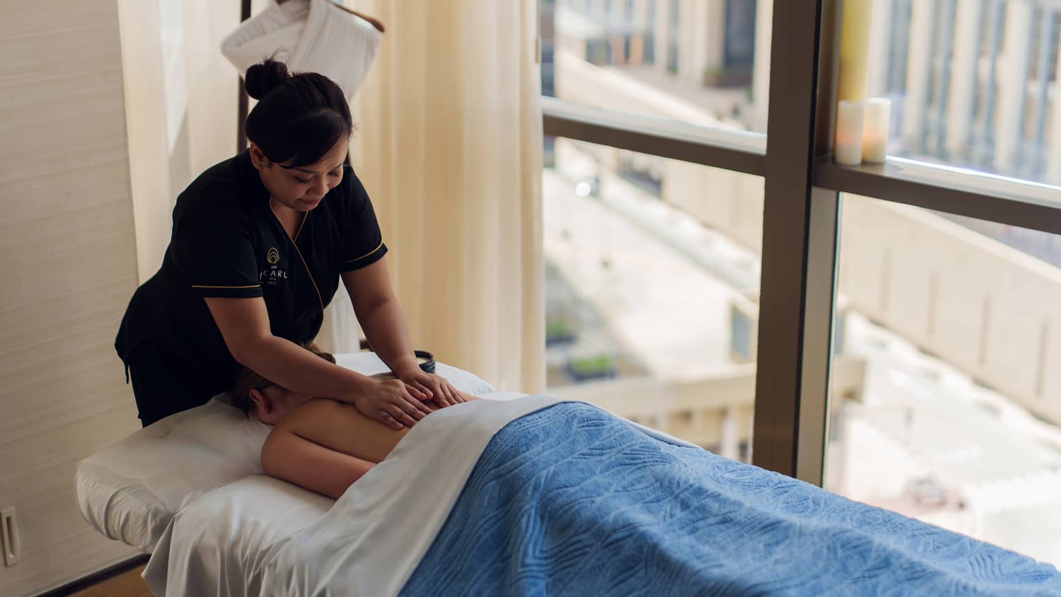 Massage therapist wearing black shirt and black pants runs their hands down the back of a guest lying face-down on a massage table beneath a white sheet and blue blanket