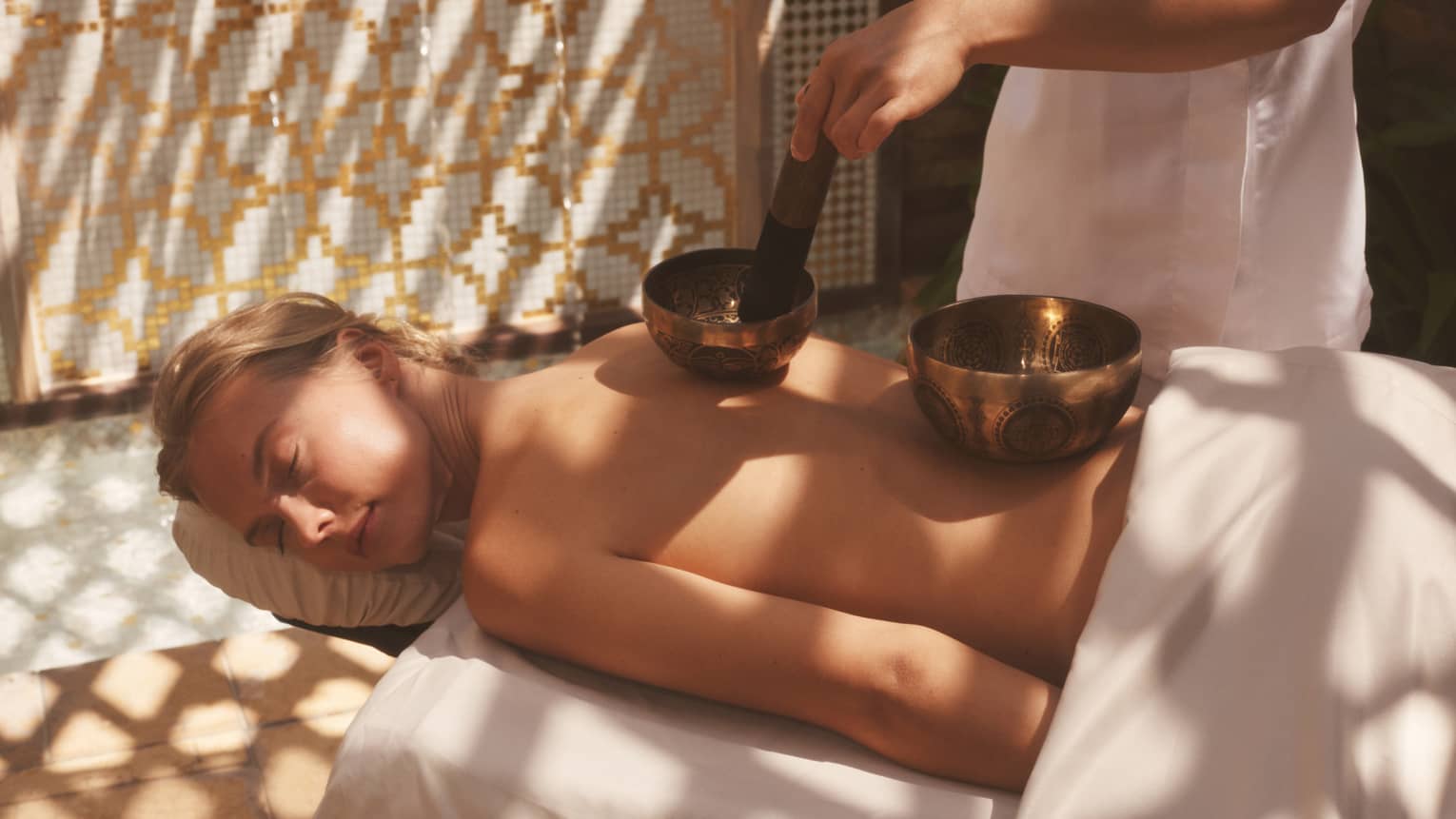 Unclothed person lying prone on a spa table with two sound healing bowls on back and spa therapist using them
