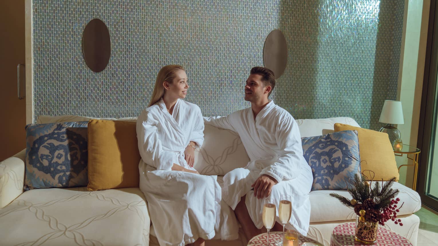 Two people each wearing a white spa robe sits on a cream-coloured couch with yellow and blue pillows in a spa waiting room