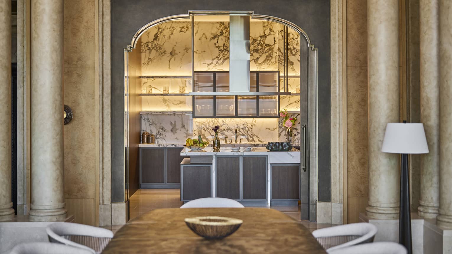 Hotel suite dining room with a wood table, white chairs, tall columns and an arched opening leading to a modern kitchen with white marble walls