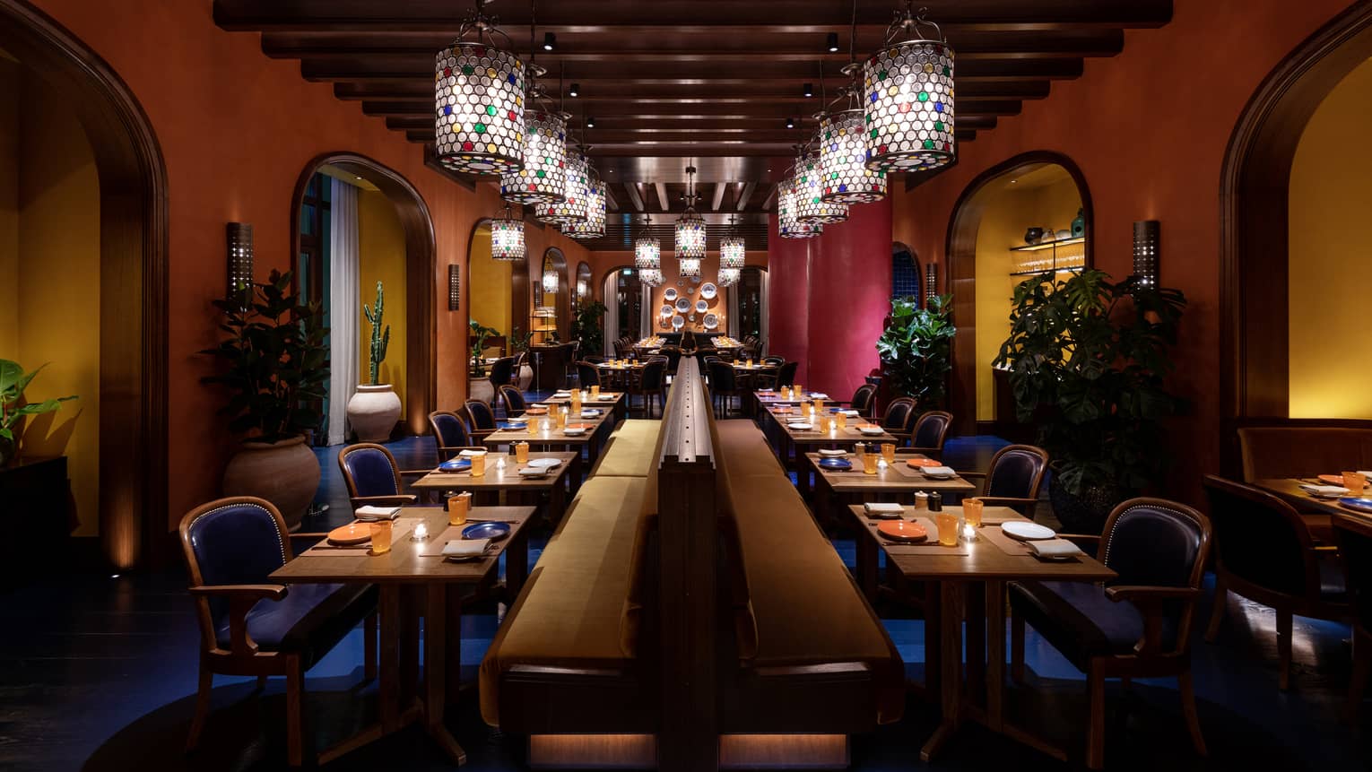 A dimly lit dining room with wood-beam ceilings and cylindrical glass chandeliers is filled with two rows of tables lining a centered banquette with rows of blue chairs on either side