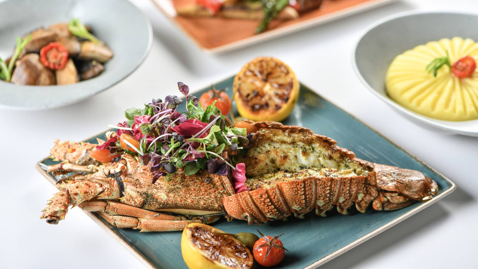 Grilled lobster on a blue square plate topped with a micro greens salad and grilled lemon halves on each side, with three small side dishes in the background