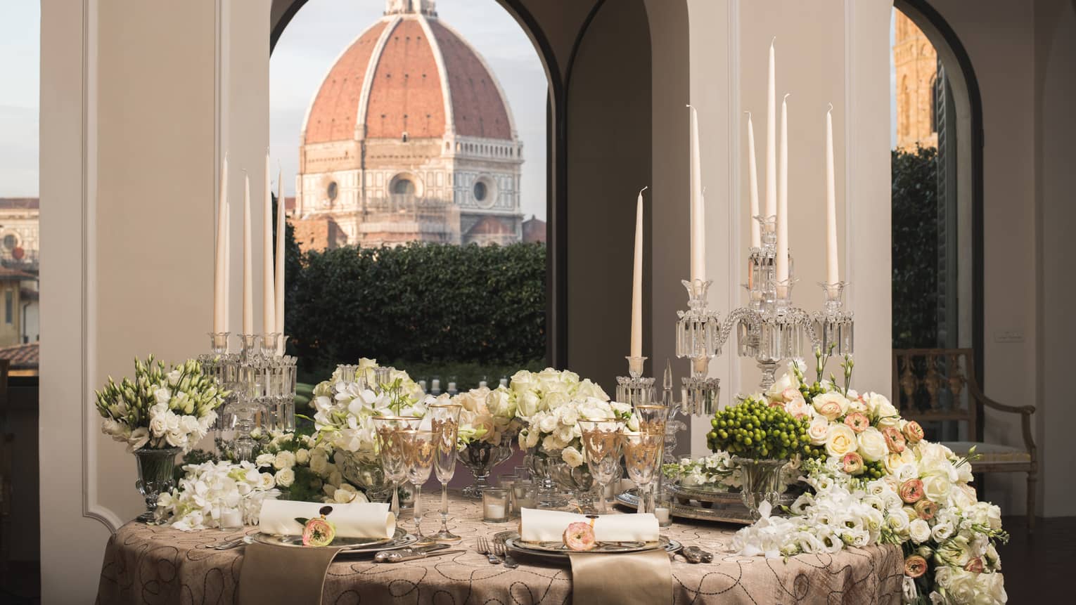 Elegant dining table set with candles, flowers, and glassware, with a large dome visible through arched windows in the background.