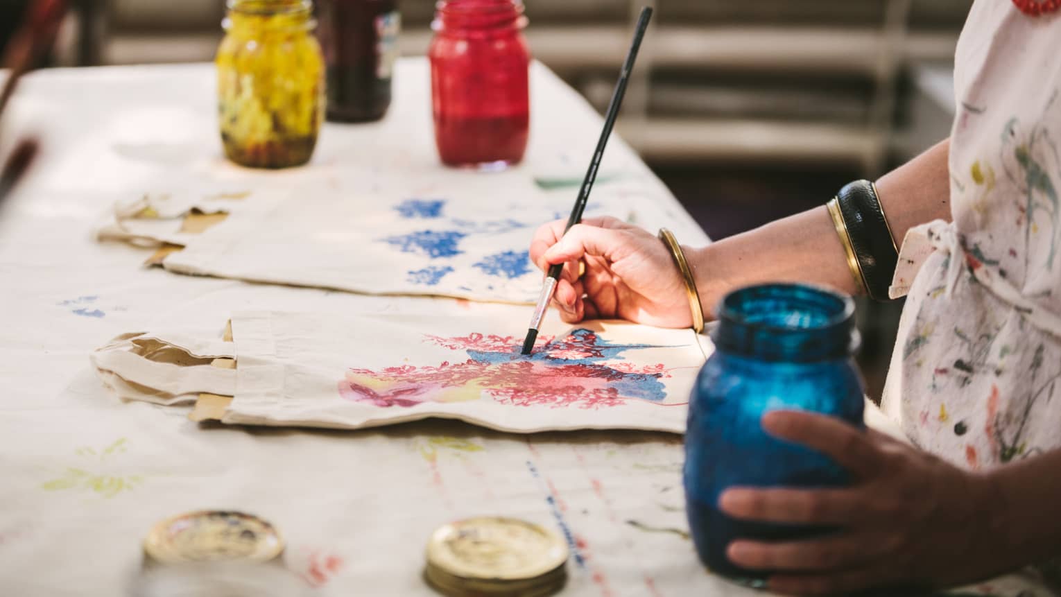 An artist using mason jars filled with red, blue, yellow and black paint to paint images of flowers on canvas tote bags.