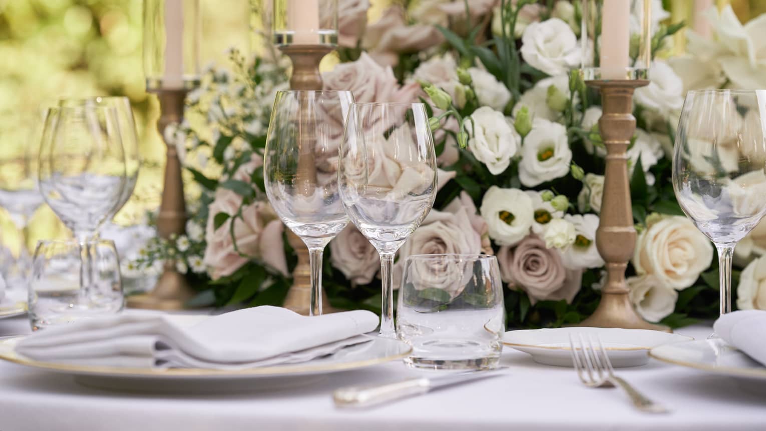 A table setting with a pale pink and white floral bouquet, gold candlesticks, wine glasses, and white plates