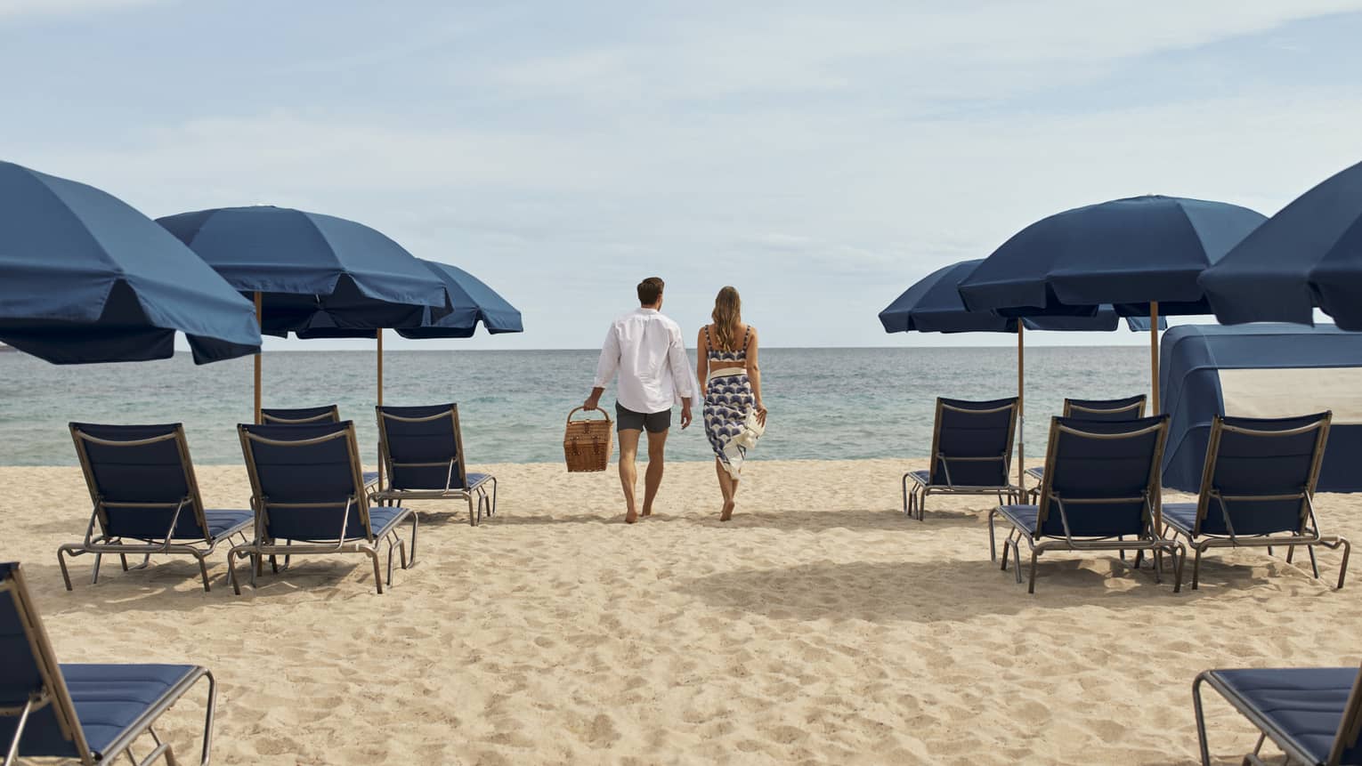 A man and woman walking between blue lounge chairs and umbrellas on sand towards the ocean.