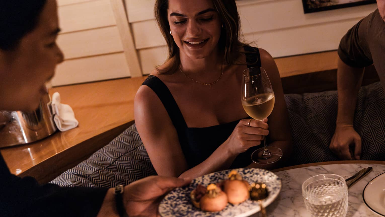 Guests sit at a booth table in a dining space. A person is handing the guests a plate of food, while the guest holds a glass of wine in a stemmed glass.