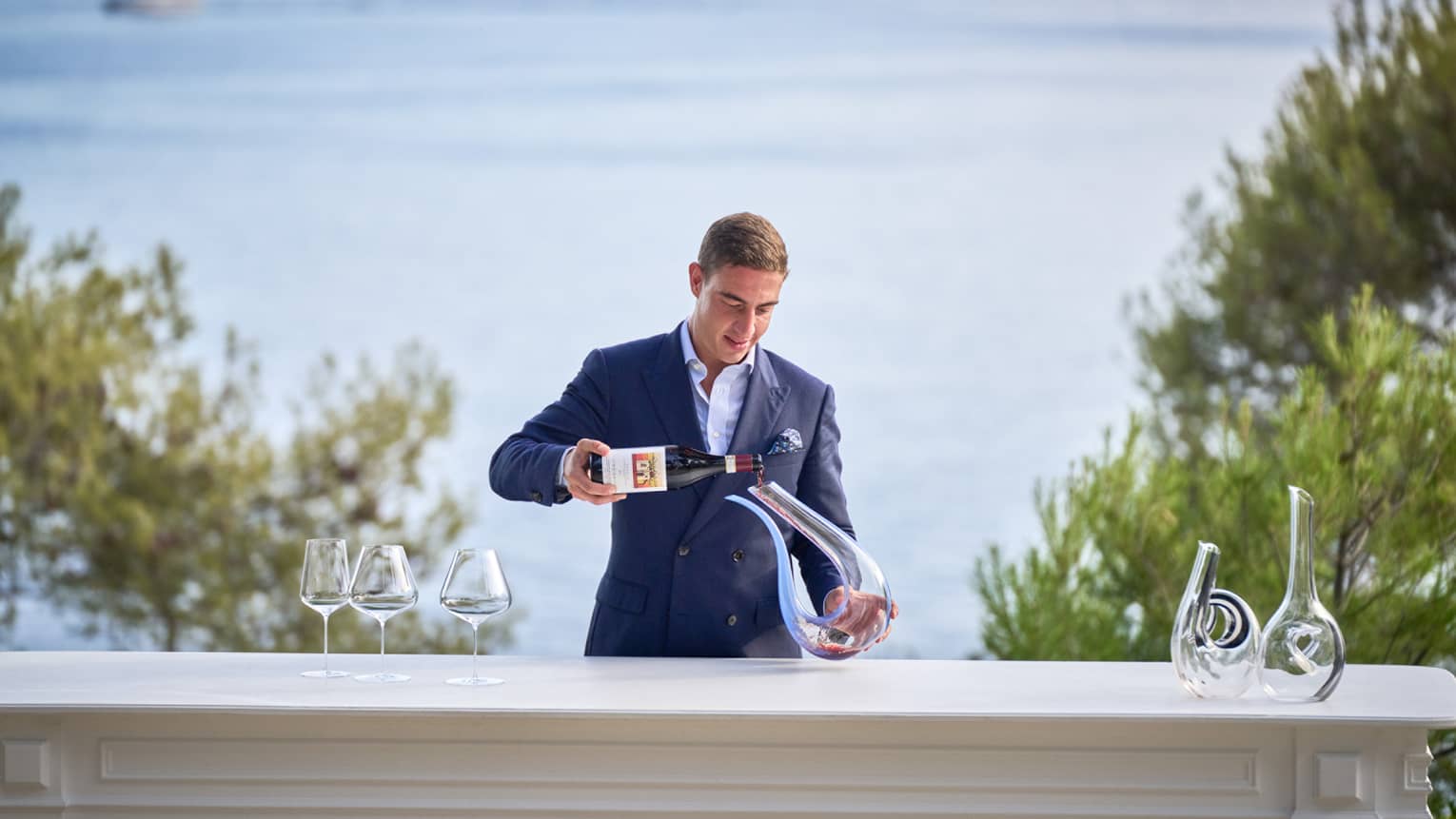 Head Sommelier Alessandro Nigro Imperiale stands behind waterfront table, decanting a bottle of red wine 