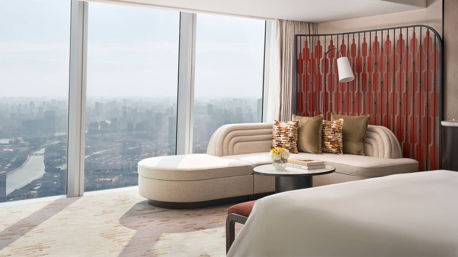 Sleek hotel room featuring a large ivory-coloured chaise next to a wall of windows overlooking city