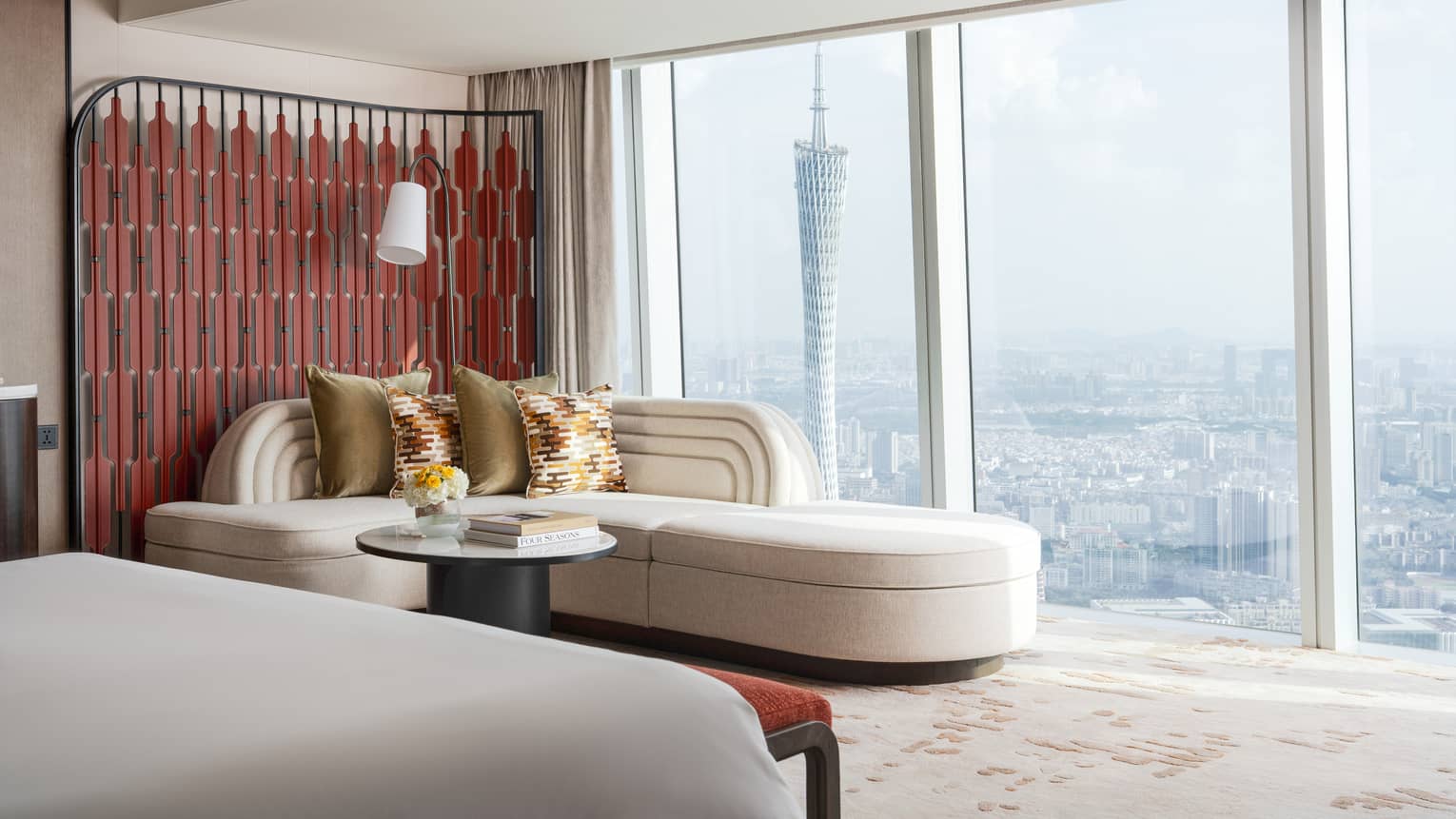 Hotel room with ivory-coloured corner sofa next to wall of windows, overlooking cityscape