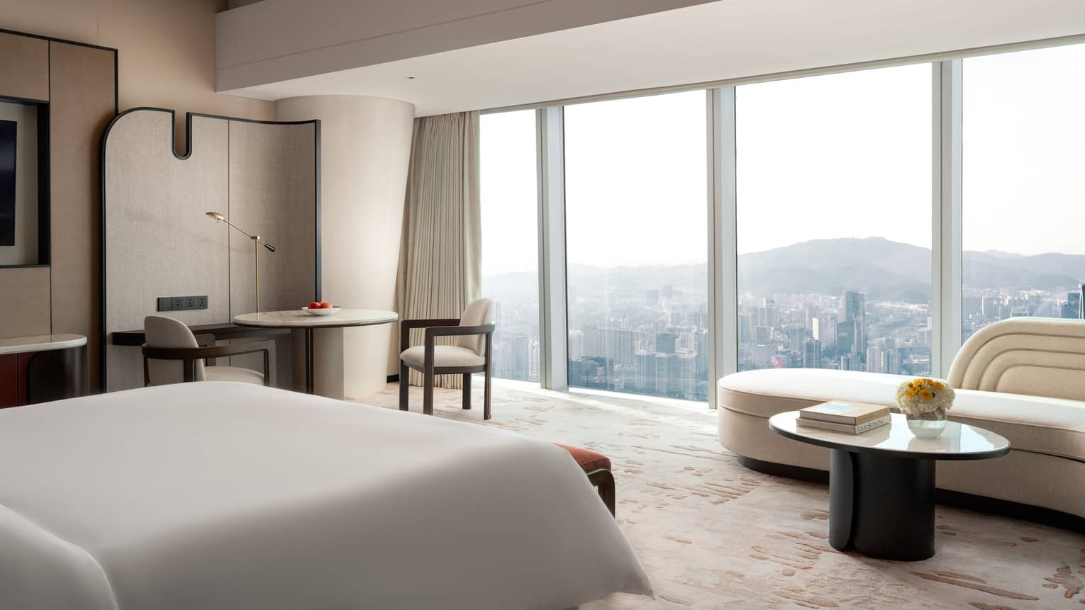 Modern hotel room with king bed, table with two chairs, and an ivory-coloured chaise next to wall of windows