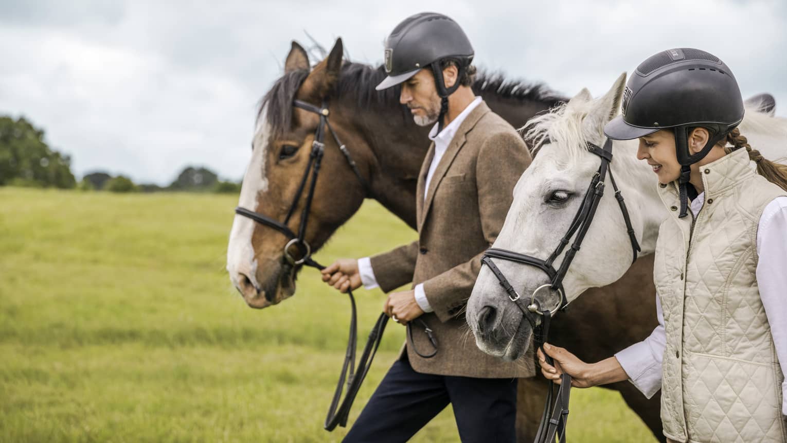 Man and woman wearing equestrian riding helmets lead horses through green field