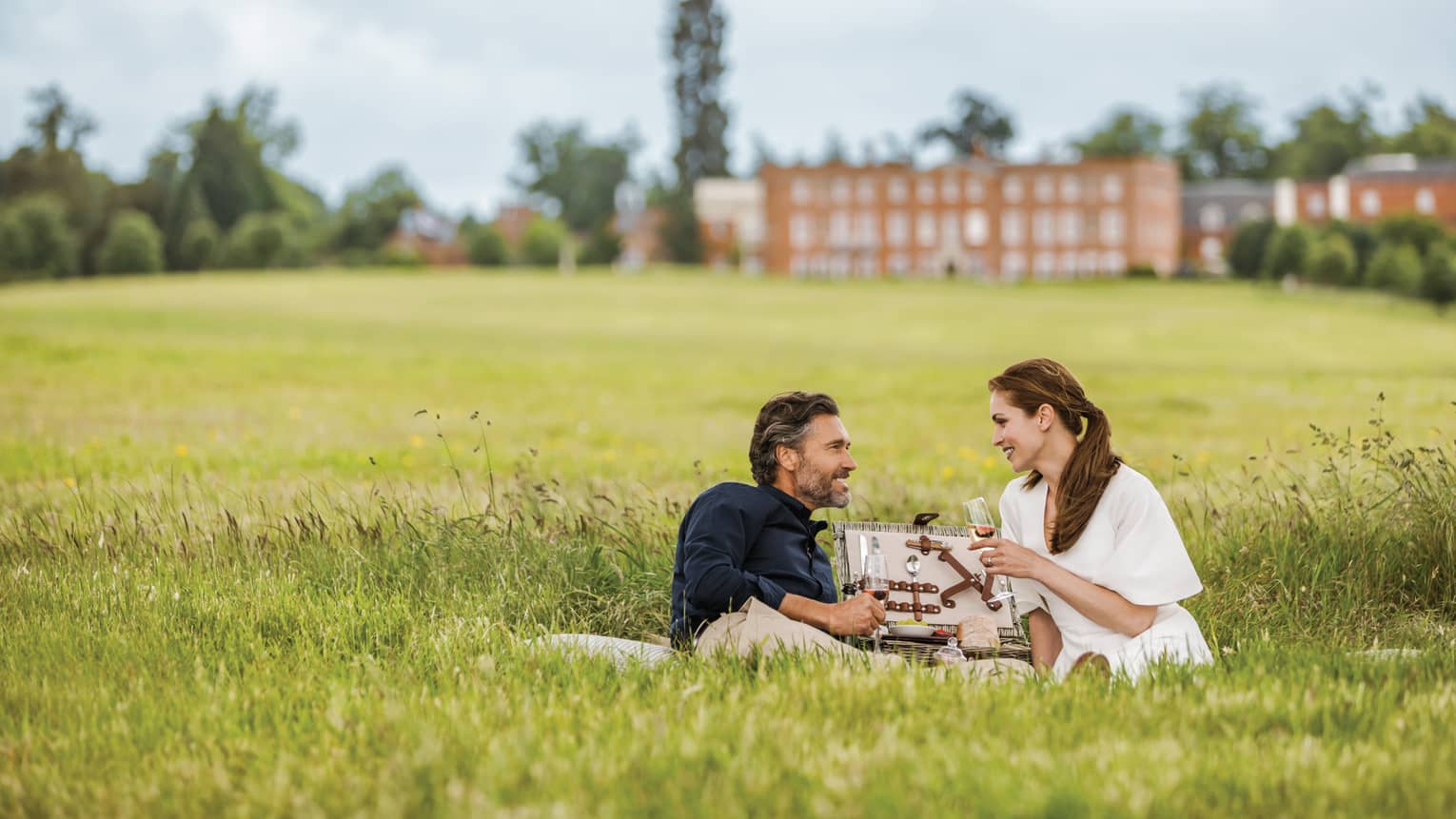 Couple with wine glasses lies in tall green grass with open picnic basket, hotel manor in background