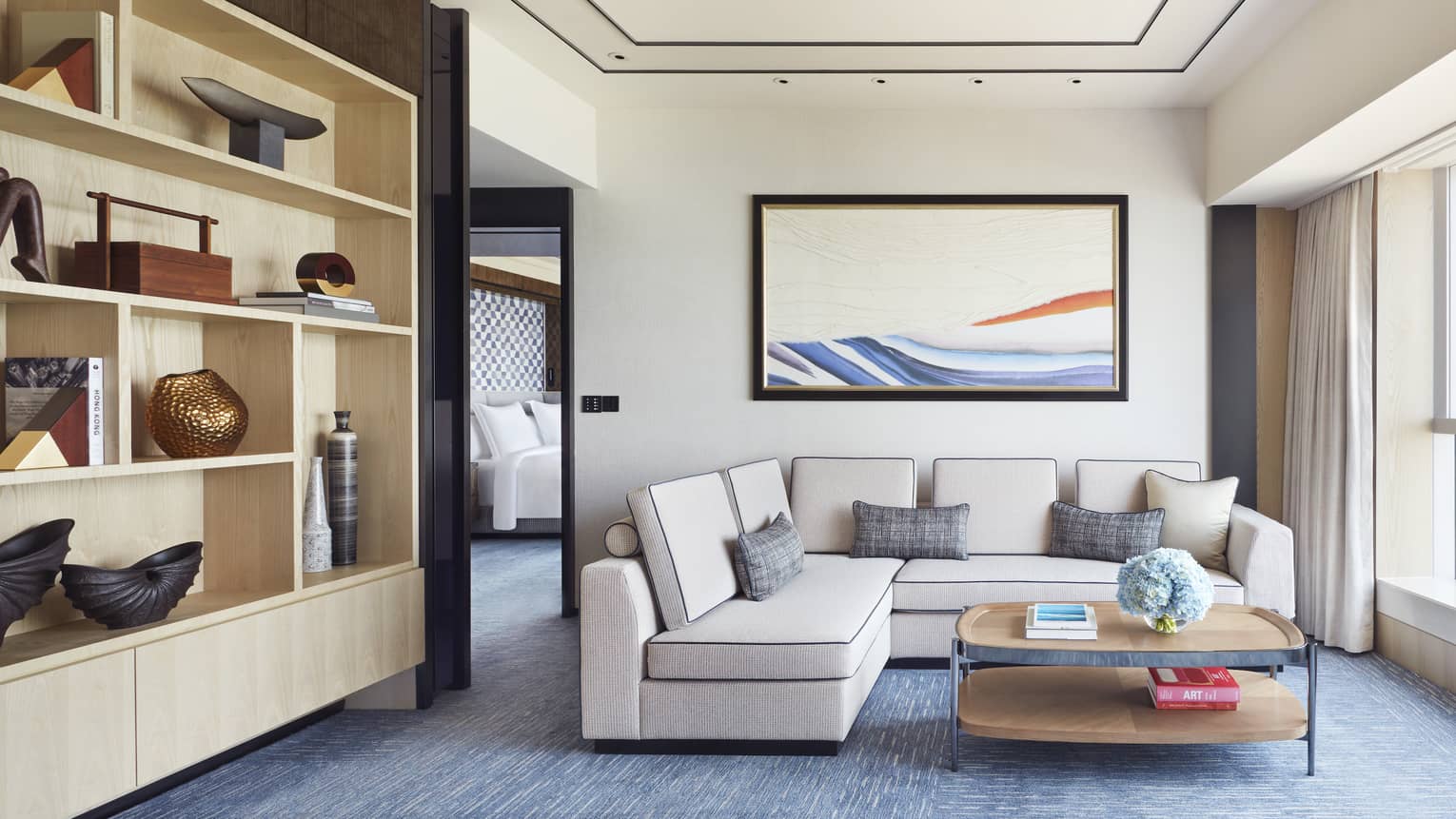 Modern hotel living area with beige sectional sofa, abstract wall art, open shelving with decor and coffee table with books and blue flowers.