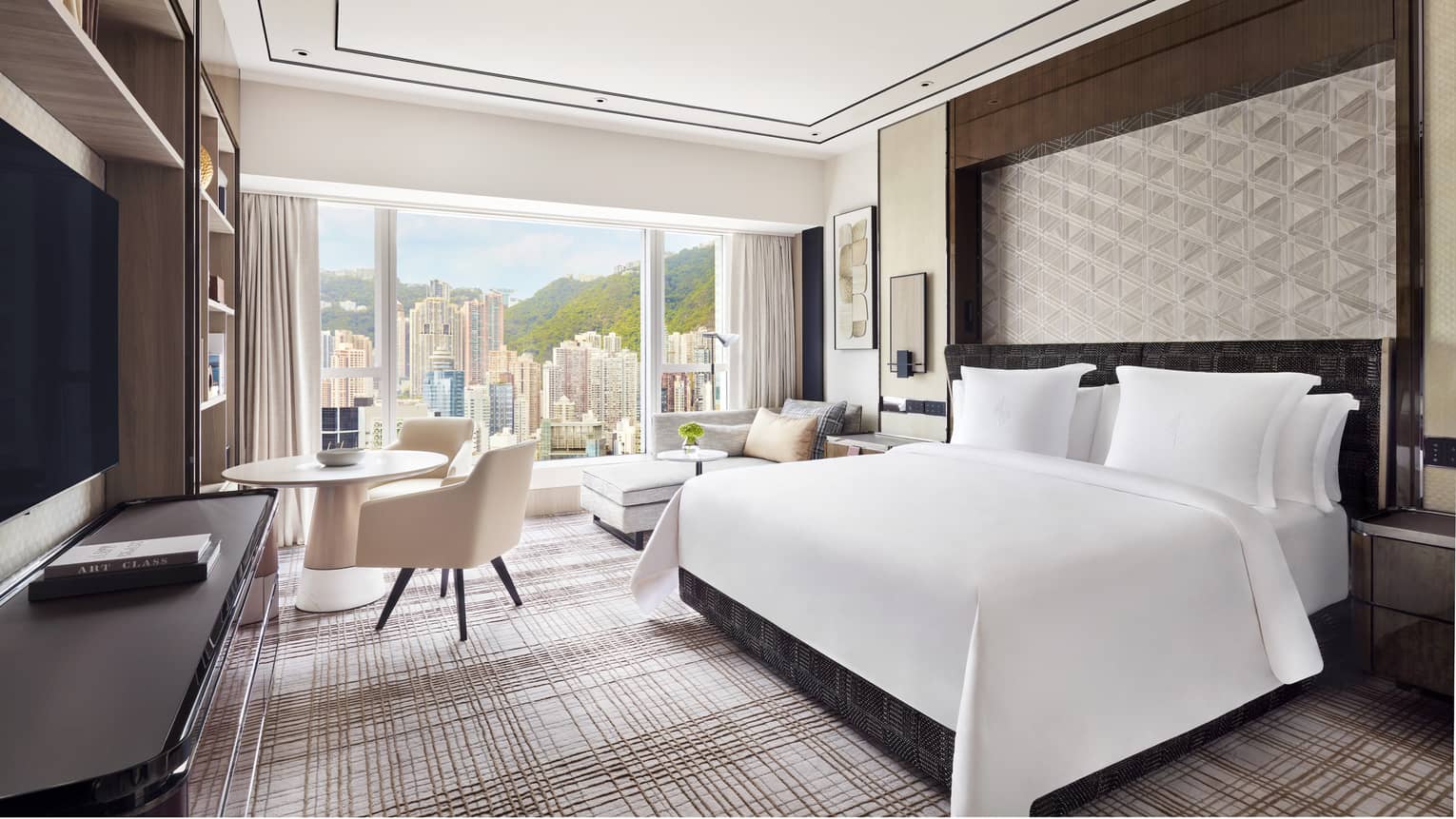 Modern hotel room with a large bed, geometric wall design, and a seating area next to a round table. Floor-to-ceiling windows offer a view of skyscrapers.