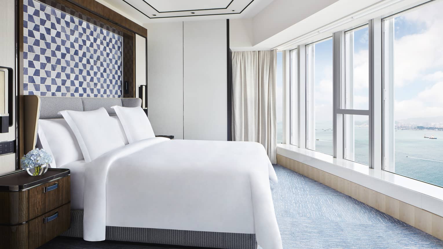Bright hotel bedroom with large bed, blue geometric headboard, floor-to-ceiling windows and ocean view.