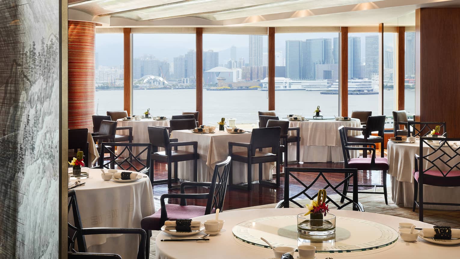 An elegant dining room with tables set with flowers and place settings, overlooking the city from across a body of water.