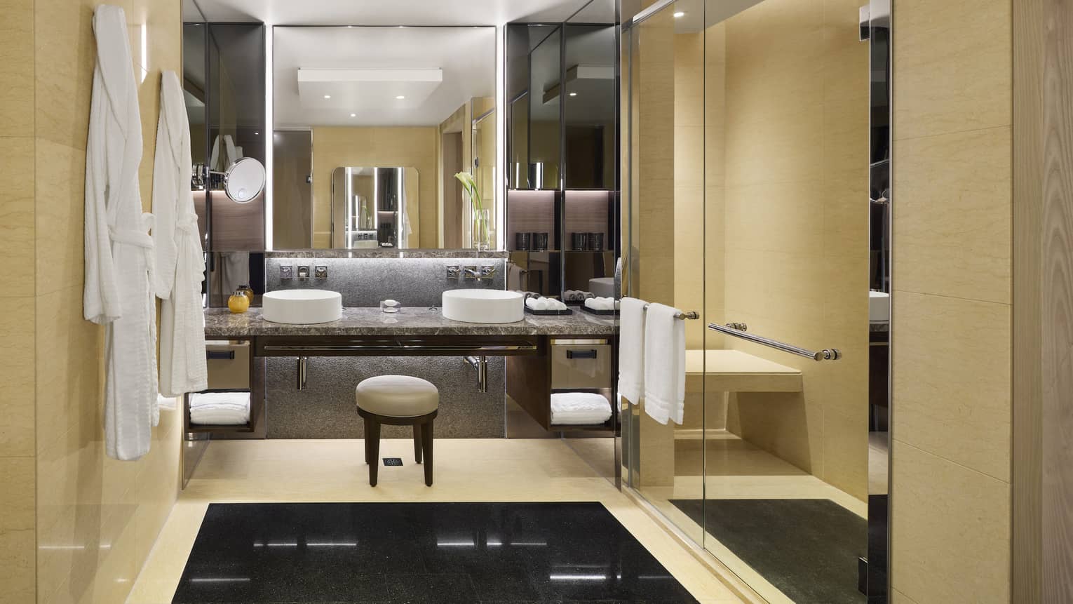Luxurious hotel bathroom with double sinks, large mirrors, beige walls, glass shower and hanging white bathrobes.