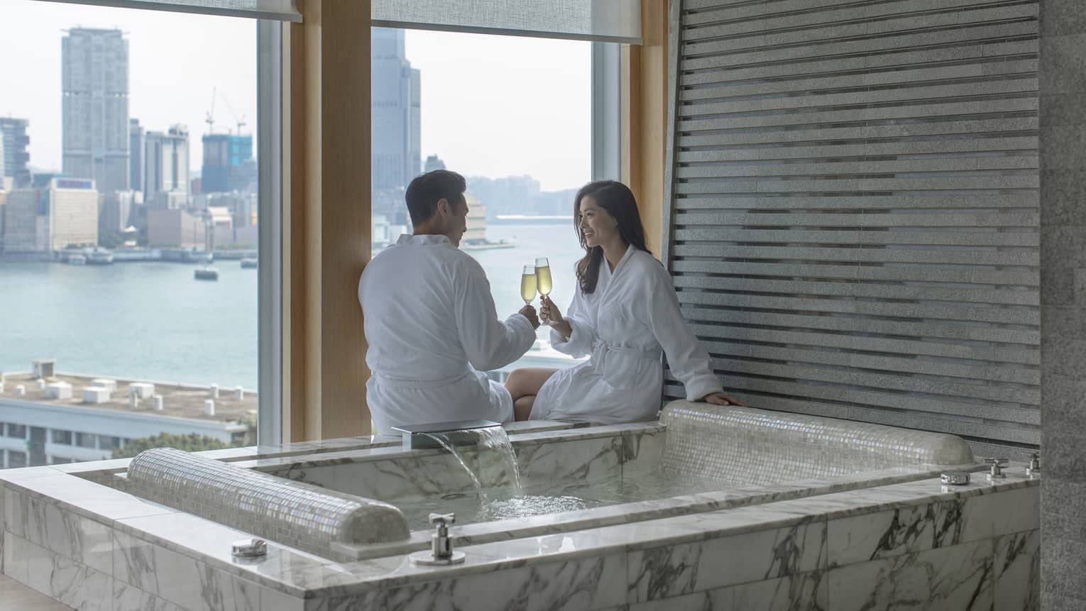 Man and woman clinking champagne glasses next to a private hot tub overlooking city harbour