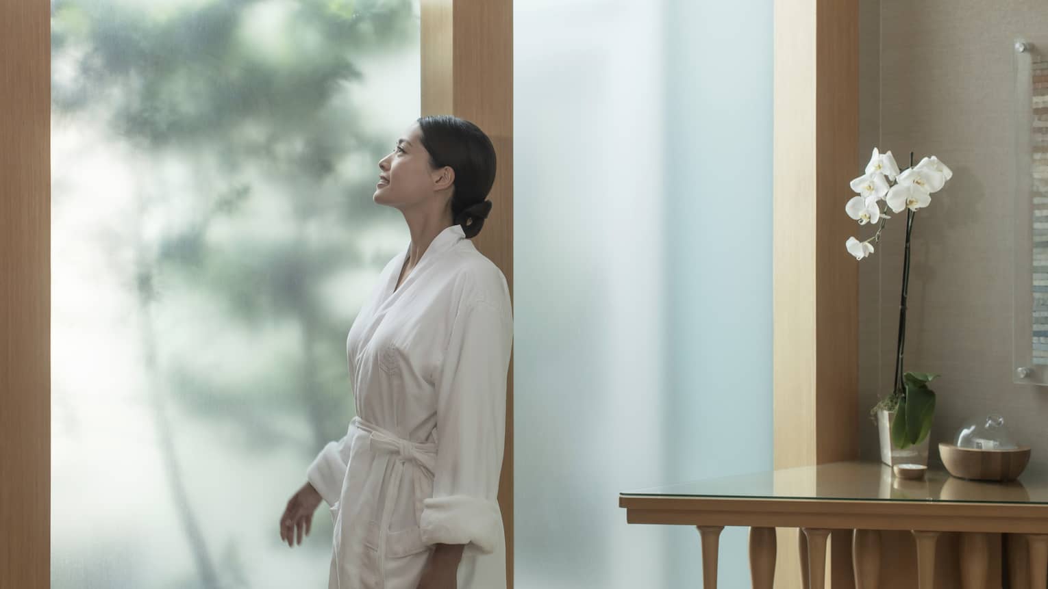 Woman wearing white robe in spa looks out frosted window