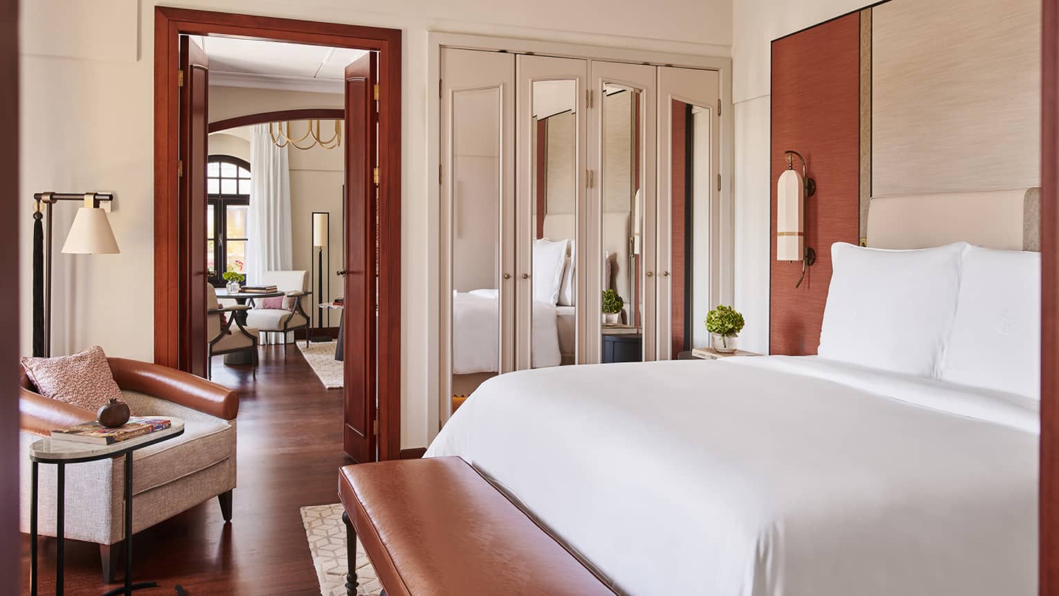 Bedroom with wooden floors, king bed, leather bench, arm chair, closet with mirrored doors and entrance into living room