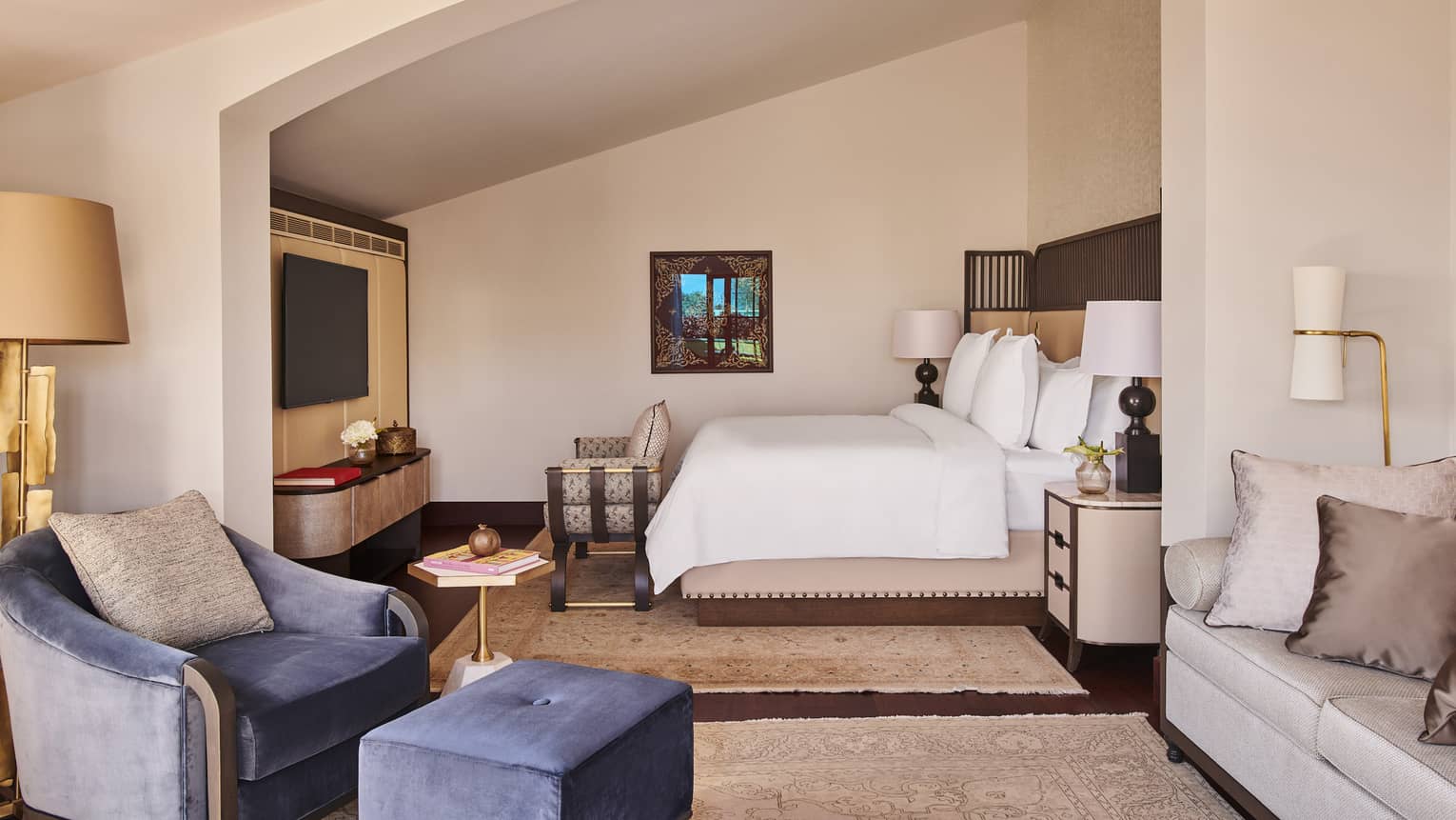 View through hotel suite’s living room to white Four Seasons bed tucked into angled alcove with chair at food and flat-screen TV on wall