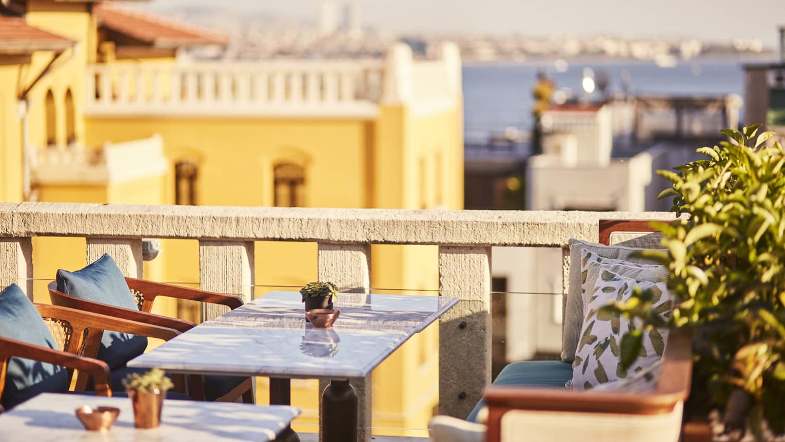 Marble table flanked by wood chairs and bench on a rooftop patio, views of beige and yellow buildings, the sea beyond. 