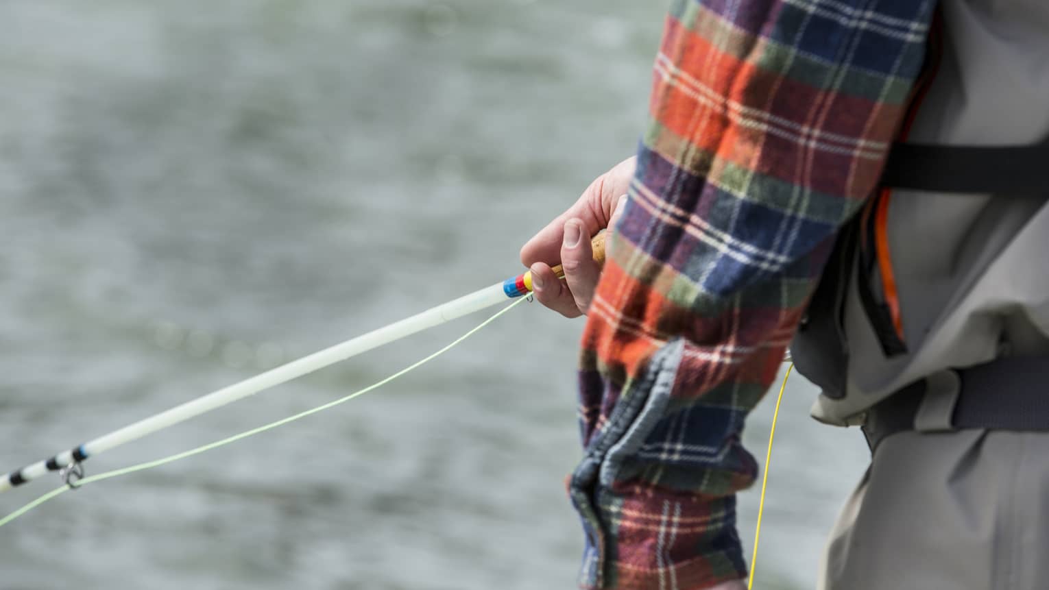 Against a background of rippling silver water, a plaid sleeve partly reveals a hand gently holding a colourful fishing rod.