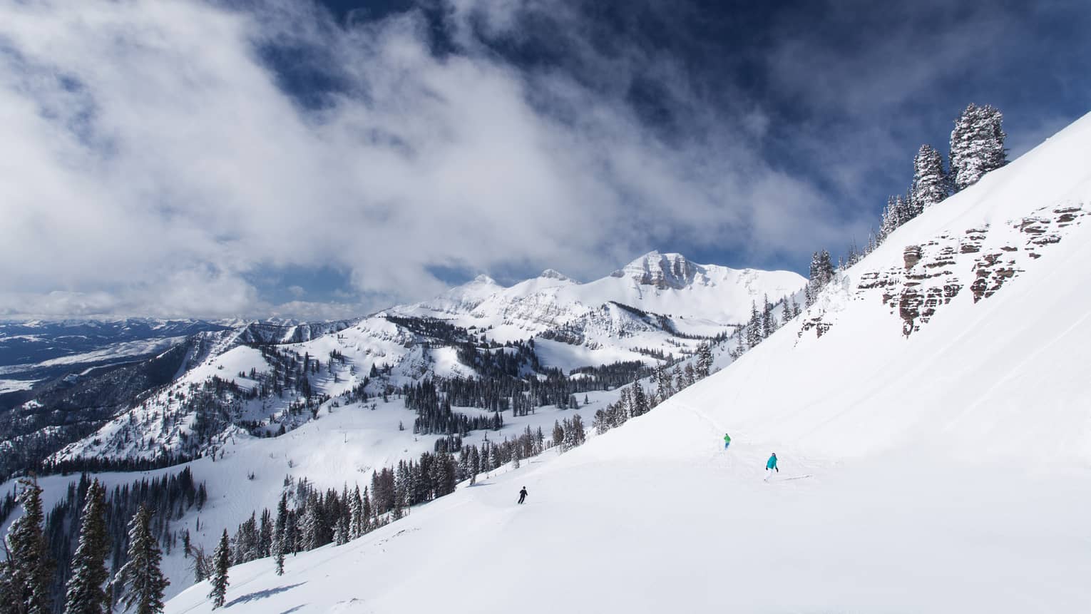 Three people ski downward, just specks of colour in the distance, the snow-capped mountains looming in the background