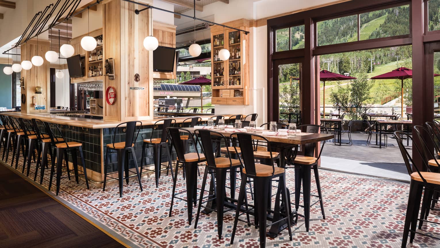 High dining tables, stools on colourful tile floors, around light wood bar, glass wall with wood trim, patio