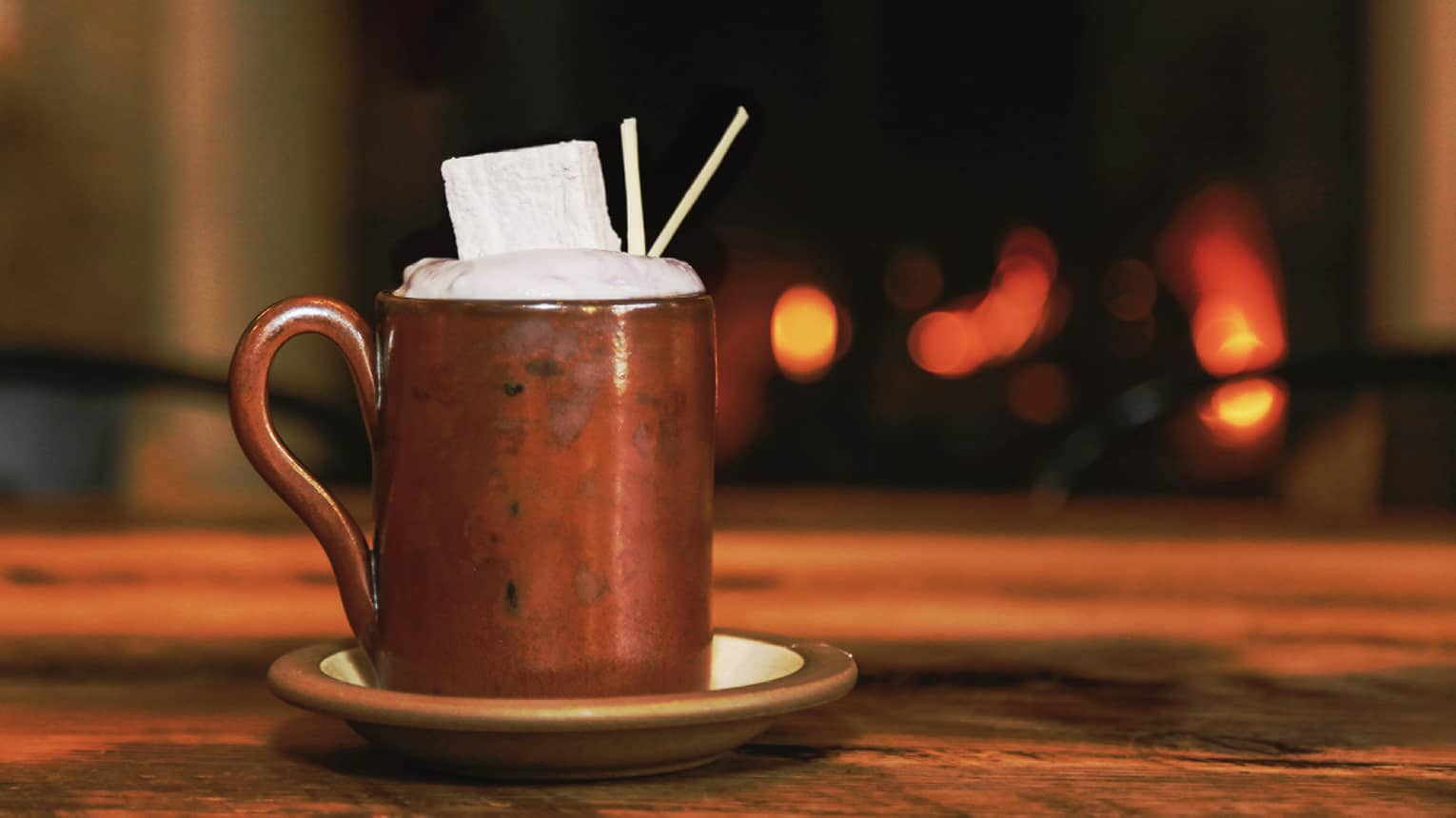 Brown mug on saucer with foamy hot cocoa, marshmallow on dark table
