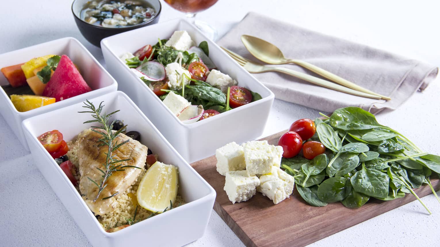 Rectangular white dishes and wood platter with salads, fruit and seared fish