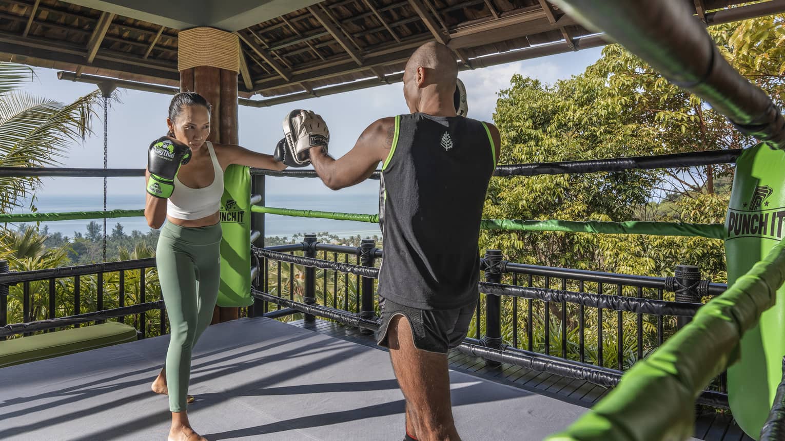 Two people practise Muay Thai in an outdoor boxing ring nestled in the treetops and overlooking the forest and the ocean.