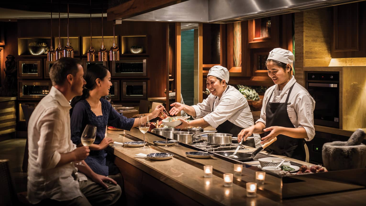 Two chefs smile and laugh with a couple as they prepare them private dinner