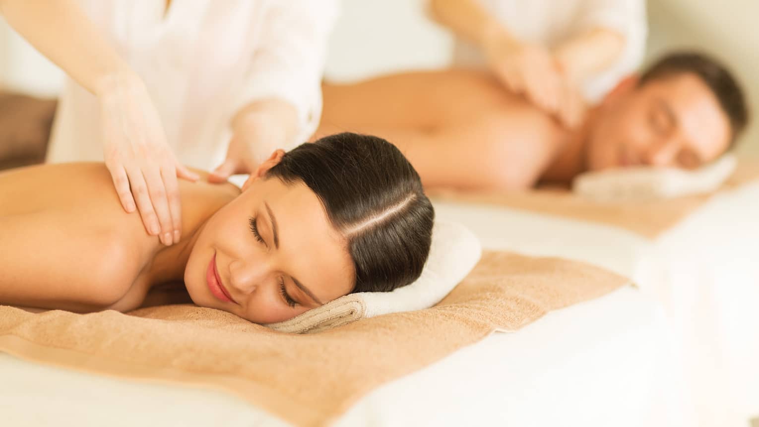 Woman and man lie side-by-side on massage tables as spa staff massage their bare shoulders