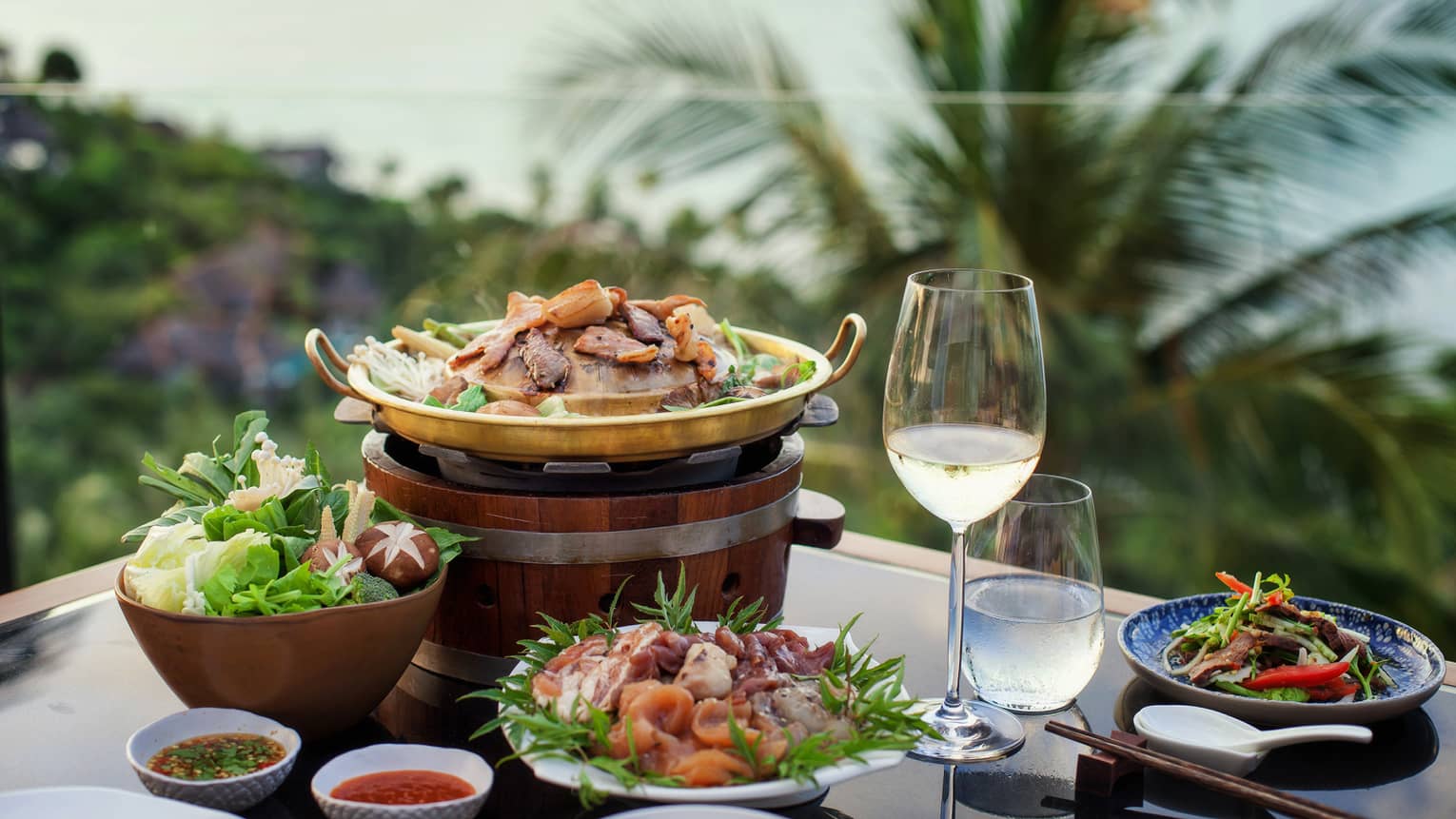 Four plates of food, glass of water and white wine, tropical scenery in background